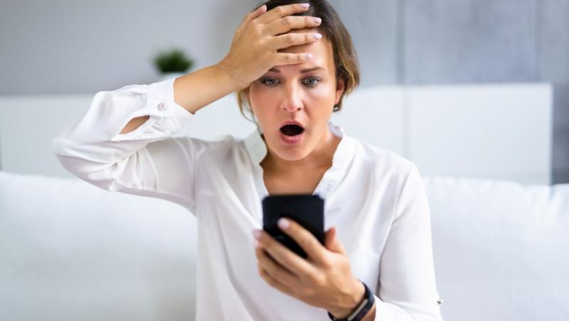 femeie, îmbrăcată într-o cămașă albă, cu telefonul într-o mână și cu cealaltă la cap, în timp ce rămâne de-a dreptul uimită de ceea ce vede în ecranul mobilului